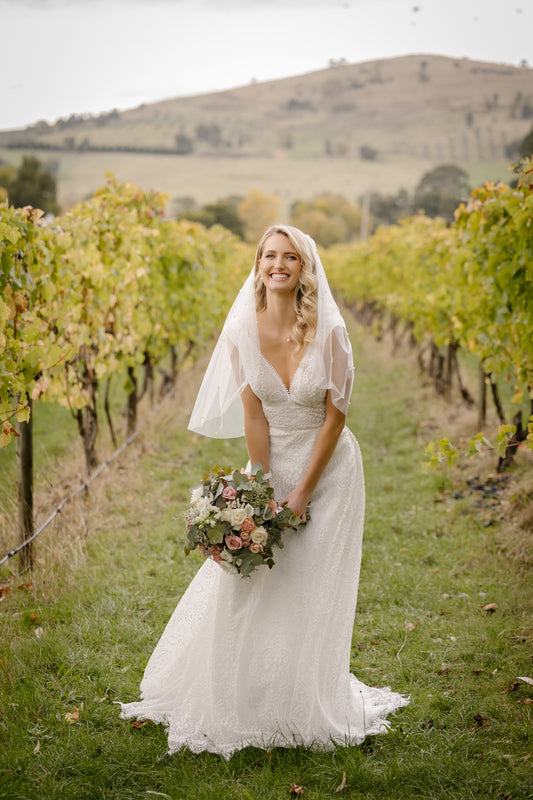 Smiling bride in a sustainable lace wedding gown, holding a bouquet, standing amidst a vineyard, reflecting Divine Bridal's commitment to eco-friendly bridal fashion.