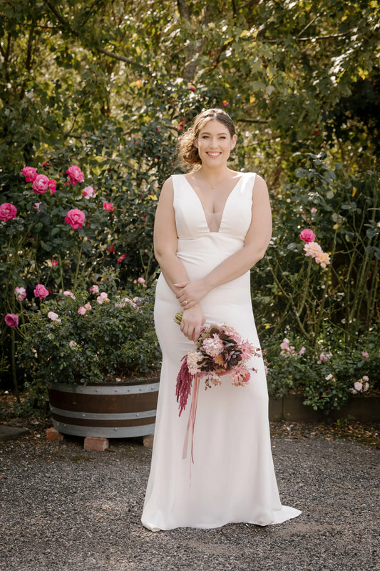 Bride in Aurora Gown – Full-Length Front View Outdoors
Bride wearing the Aurora wedding dress featuring thin straps, a plunging neckline with mesh insert, and a sleek crêpe skirt that contours the body.