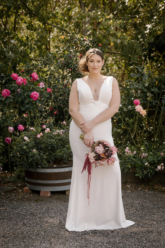 Aurora Plunge Neckline Bridal Gown – Full View in Garden Setting
Elegant full-length view of bride in fitted crêpe Aurora gown with a dramatic plunge neckline and delicate straps, surrounded by greenery.