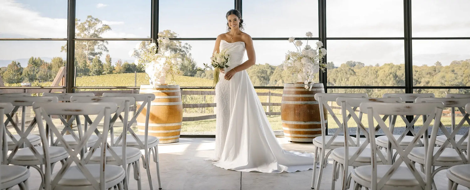 Bride wearing the Nina luxury wedding dress with detachable satin overskirt in a modern winery setting, featuring a strapless bodice and flowing chapel train.