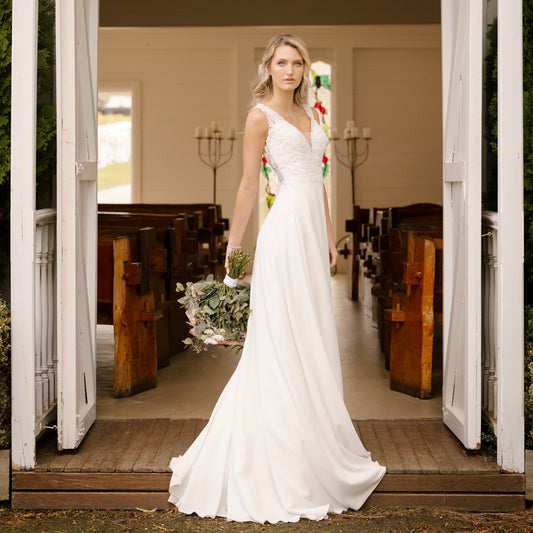 An elegant bride stands at the entrance of a charming chapel, poised in the Luna Wedding Gown. The dress features a plunging V-neckline with illusion mesh and form-fitting silhouette. The gown's smooth fabric cascades into a graceful chapel train, while the bride holds a bouquet of soft-colored flowers, completing her timeless bridal look.