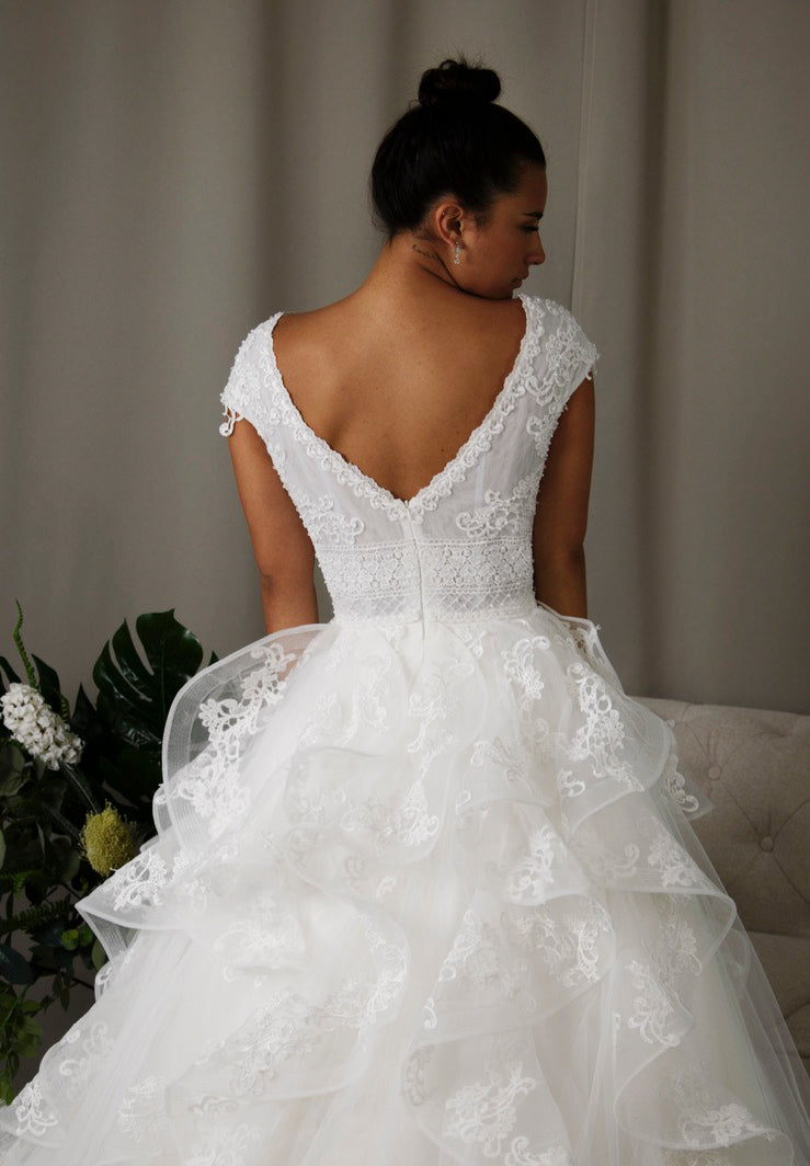 Bride wearing Lucille wedding dress, a stunning strap V-neckline lace ball gown with intricate beadwork, a tiered tulle skirt, and chic back design.