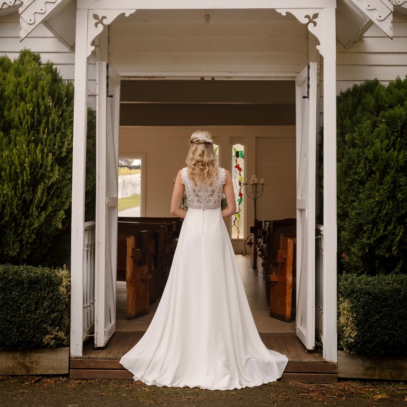 An elegant bride stands at the entrance of a charming chapel, poised in the Luna Wedding Gown. The dress features a plunging V-neckline with illusion mesh and form-fitting silhouette. The gown's smooth fabric cascades into a graceful chapel train, while the bride holds a bouquet of soft-colored flowers, completing her timeless bridal look.