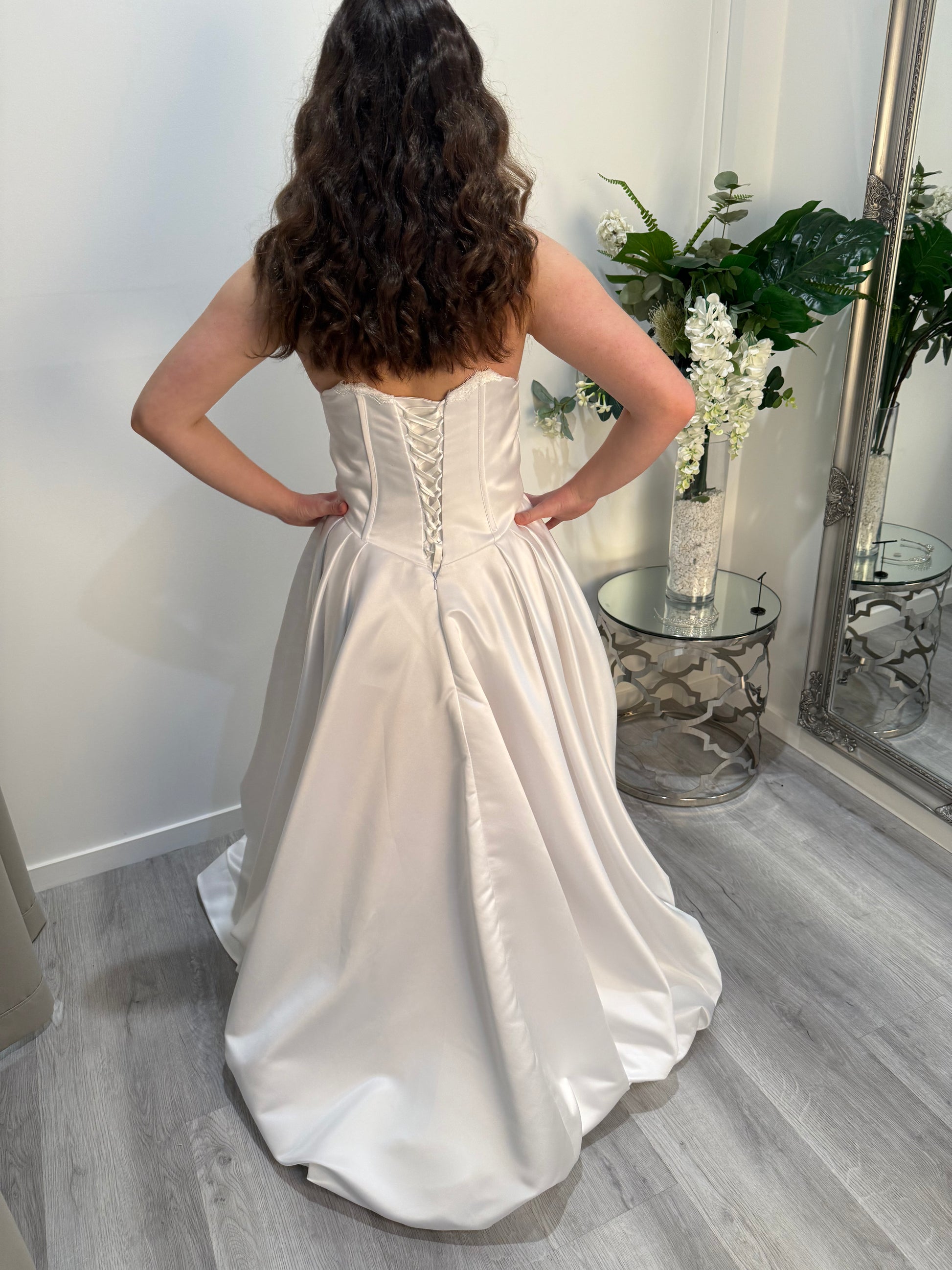 Woman wearing a white strapless gown standing in a room with a mirror and flowers.