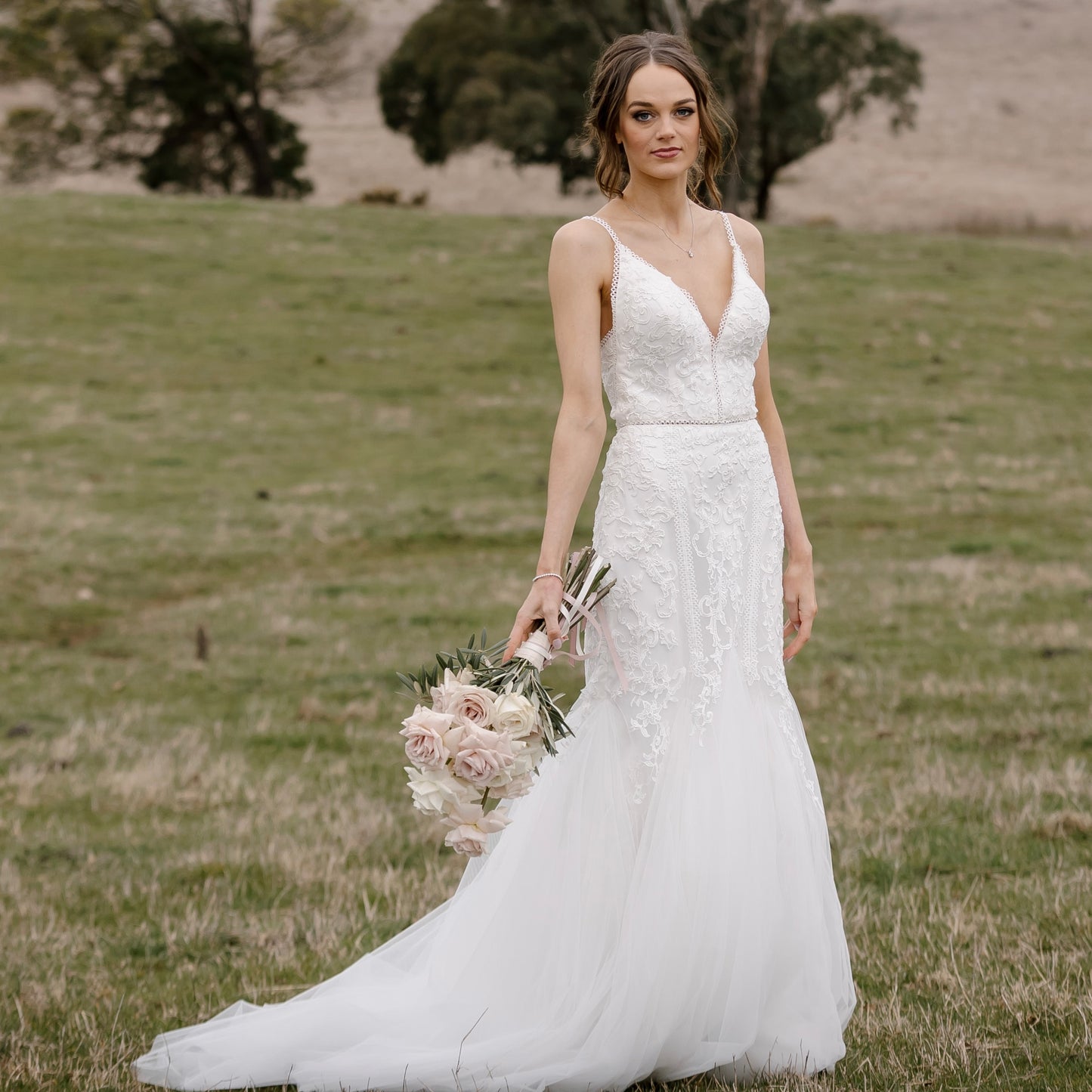 "A captivating and elegant Selena wedding gown, featuring a Boho-inspired design. The dress showcases a flattering V-neckline with delicate lace trim, intricate lace detailing throughout, and a fit and flare silhouette that accentuates the wearer's curves. The gown is completed with a chapel train and lace trim on the waist, creating a stunning bridal look."