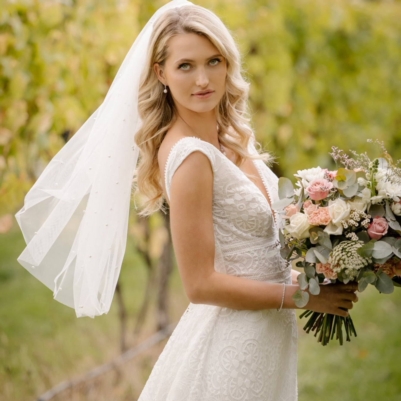 A radiant bride in the Tiana Wedding Gown smiles joyfully amidst a vineyard setting. The gown features an intricate full-lace overlay, scalloped neckline, and a romantic train, complemented by a delicate veil and a bouquet of soft roses and greenery