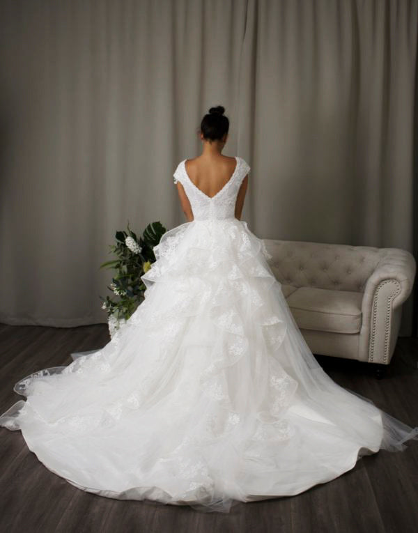 Bride wearing Lucille wedding dress, a stunning strap V-neckline lace ball gown with intricate beadwork, a tiered tulle skirt, and chic back design.