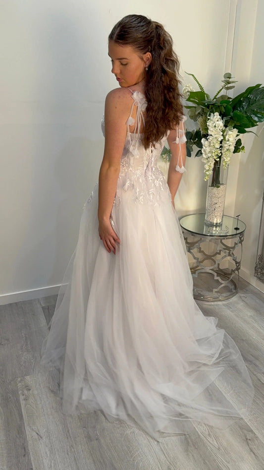 Woman in a white floral dress standing in a room with a vase and flowers in the background.