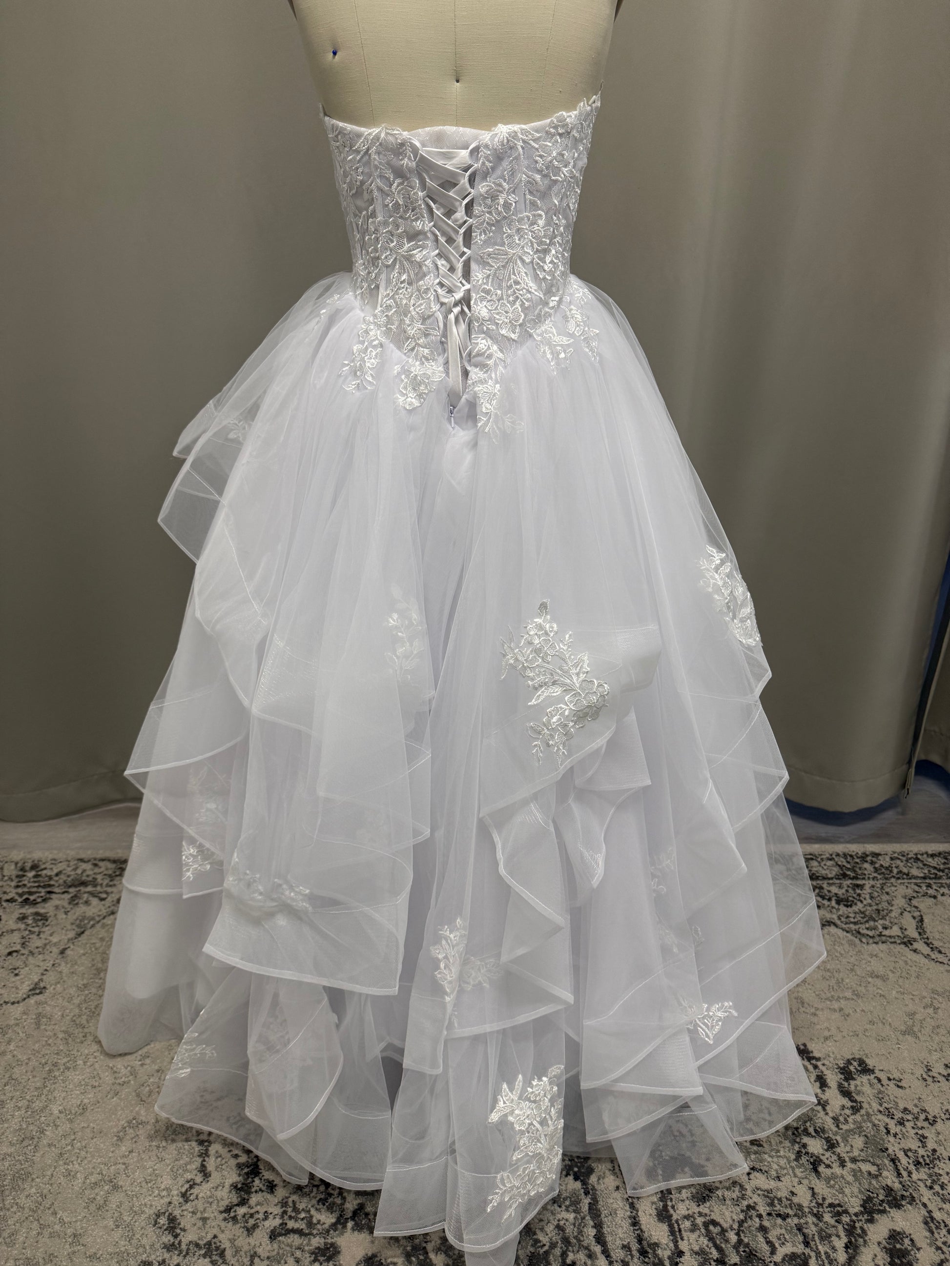 Kasey V1 lace appliqué debutante dress back view showing lace-up corset, floral lace and full white ruffle skirt with scattered lace
