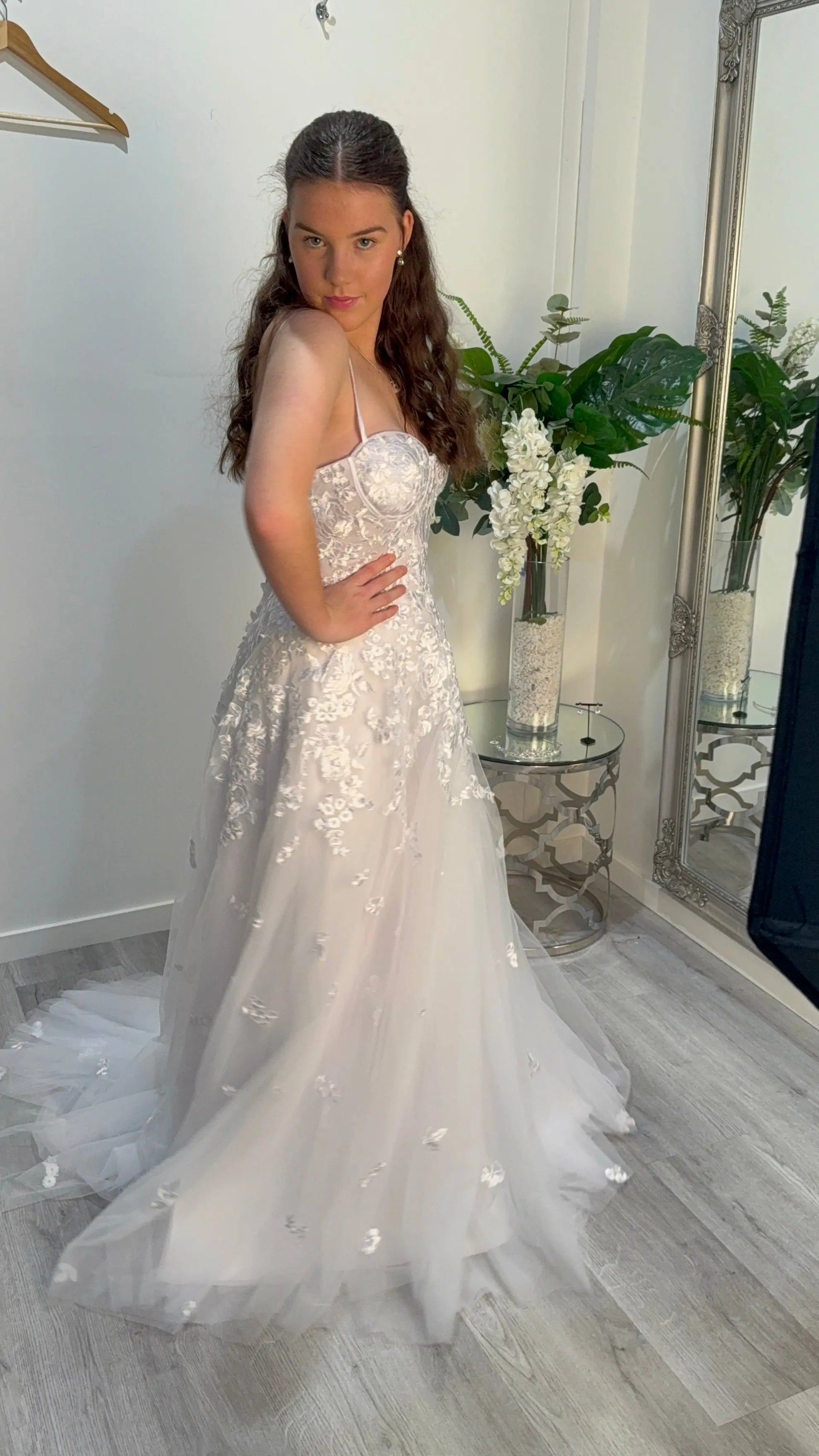 Woman in a white debutante dress standing in a room with a mirror and plants.